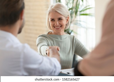 Head shot happy mature businesswoman shaking hands with partners. Smiling middle aged female lawyer, financial advisor or real estate agent making agreement with diverse family couple clients.