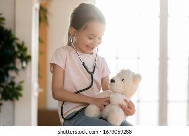 Head Shot Happy Playful Little Patient Wearing Stethoscope, Pretending To Be Doctor Checking Lungs Of Fluffy Toy In Clinic Or Home. Happy Small Girl Playing Nurse, Holding Favorite Teddy Dog.