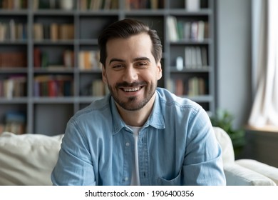 Head shot happy young 30s handsome caucasian man sitting on sofa, looking at camera, involved in web camera conversation with family or friends, enjoying video call distant communication at home.