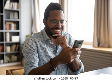 Head shot happy young african american man in eyeglasses looking in phone screen. Smiling millennial biracial guy reading pleasant good news sms in smartphone, enjoying chatting communicating online.