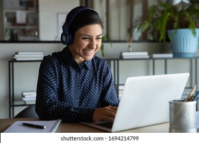 Head shot happy young indian ethnicity female manager wearing wireless headphones, looking at laptop screen, holding pleasant conversation with partners clients online, working remotely at workplace. - Shutterstock ID 1694214799
