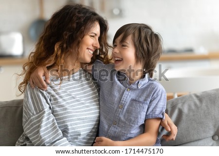 Head shot happy young woman cuddling talking to small kid son, having fun together on couch in studio living room. Smiling child boy communicating with elder sister nanny babysitter, laughing at home.