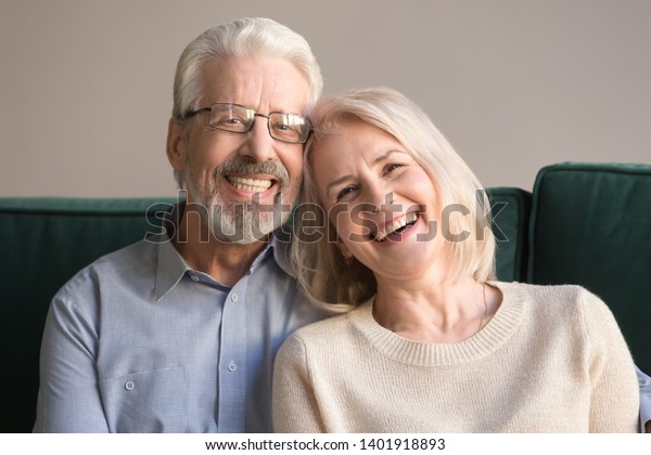 Head shot laughing retirees pretty couple sitting
on couch at home, spouses having candid healthy toothy smile,
dental treatment check-up services for old people, medical
insurance health care
concept
