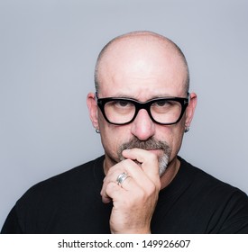 Head Shot Of A Middle Aged Bald Man With A Goatee, Black Glasses And A Black Shirt Looking At The Camera In Deep Thought With His Hand To His Face