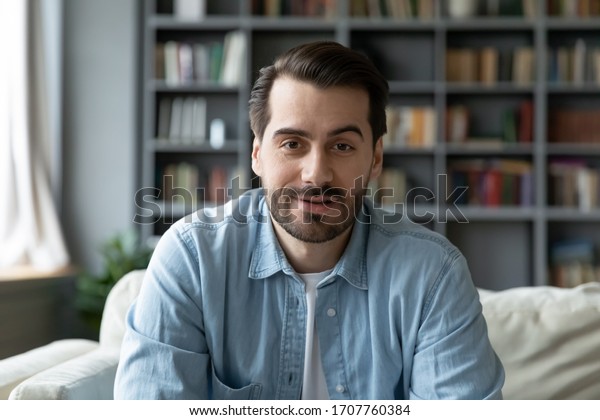 Head shot millennial guy sit on sofa in living\
room makes video call looks at camera, conversation by distant\
videocall, distance hiring job interview process, tutor and trainee\
study on-line concept