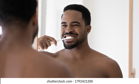 Head shot mirror reflection African American handsome young man with healthy toothy smile cleaning brushing teeth, standing in bathroom, enjoying morning routine, oral hygiene concept - Powered by Shutterstock