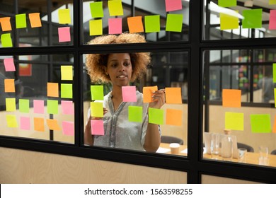 Head shot pleasant concentrated millennial african american female manager planning project workflow, putting paper stickers with tasks on window glass kanban brainstorming board at modern office.