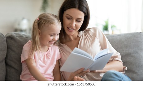 Head Shot Pleasant Young Mother Sitting On Couch With Adorable Kid Girl, Reading Fairy Tales Together. Involved Small Daughter Looking In Book With Interesting Stories, Enjoying Relaxing Time At Home.