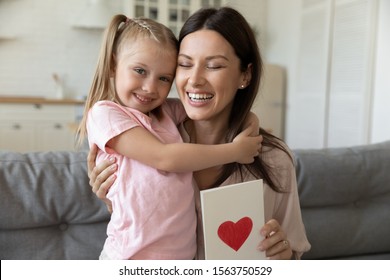 Head Shot Portrait Adorable Smiling Cute Small Girl Embracing Happy 30s Mommy With Greeting Love Card In Hands. Little Daughter Congratulating Joyful Proud Mom With Birthday Or Mother S Day At Home.