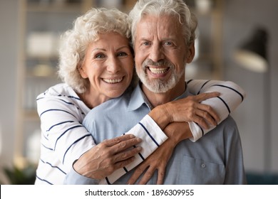 Head Shot Portrait Of Affectionate Loving Middle Aged Hoary Beautiful Woman Cuddling From Back Smiling Old Husband. Happy Loving Mature Married Family Couple Looking At Camera, Posing For Photo.