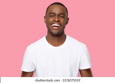Head shot portrait African American man with beaming wide healthy smile looking at camera, handsome laughing young male, funny cheerful guy in white t-shirt, isolated on pink studio background