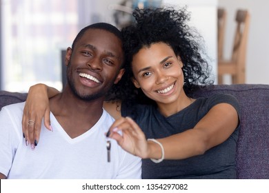 Head shot portrait african mixed race couple sitting on couch embracing feels happy holding keys from new modern first house smiling looking at camera. Move day, buy real estate loan mortgage concept