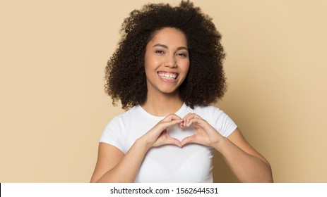 Head shot portrait beautiful African American girl with toothy smile showing heart gesture, happy young female wearing white t-shirt looking at camera, standing isolated on brown background - Shutterstock ID 1562644531