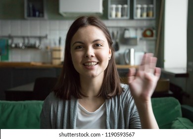 Head Shot Portrait Of Cheerful Young Woman Sitting On Sofa At Home. Girl In Casual Clothes Have Video Call Using Computer, Wave Her Hand Greeting Or Say Goodbye To Friend And Smiling Looking At Camera