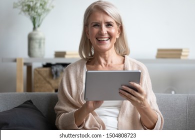 Head Shot Portrait Close Up Happy Mature Woman Using Computer Tablet, Looking At Camera, Laughing At Funny Joke Or Video In Social Network, Older Female Holding Electronic Device, Sitting On Couch