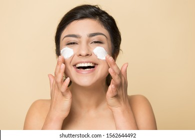 Head Shot Portrait Close Up Happy Indian Woman With Perfect Skin And Healthy Toothy Smile Applying Face Cream, Moisturizer On Cheeks, Isolated On Brown Background, Skincare And Treatment Concept