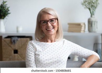 Head Shot Portrait Close Up Positive Satisfied Mature Woman In Glasses Sitting On Sofa At Home, Happy Older Female With Toothy Smile Looking At Camera, Posing For Photo On Comfortable Couch