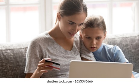 Head Shot Portrait Concentrated Young Mom With School Cute Daughter Sitting On Couch With Laptop, Making Purchases In Popular Internet Store. Smiling Woman Holding Credit Card For Online Payments.