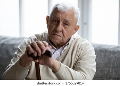 Head shot portrait depressed lonely elderly man with wooden cane looking at camera, frustrated mature male folded hands on walking stick, sitting on couch alone, loneliness and solitude