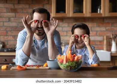 Head Shot Portrait Father And Son Having Fun With Tomatoes, Making Funny Faces, Sitting At Wooden Table In Kitchen At Home, Happy Dad With 8s Kid Child Cooking Salad, Enjoying Leisure Time Together