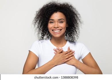 Head shot portrait happy african woman in white t-shirt pose grey background smiling looking at camera holding hands on chest feels gratitude, gesture of sincere feelings from heart and love concept