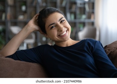 Head shot portrait happy beautiful young Indian ethnicity female homeowner resting on cozy sofa, looking at camera. Pleasant woman showing perfect whitening smile, holding video call at home.