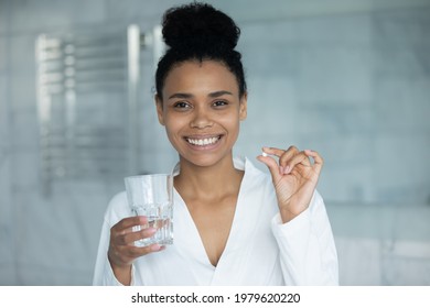 Head shot portrait of happy mixed race woman in bathrobe taking tablet, pill of capsule of vitamin, food supplement, medication, holding glass of water, smiling at camera. Healthcare, pharmacy concept - Powered by Shutterstock