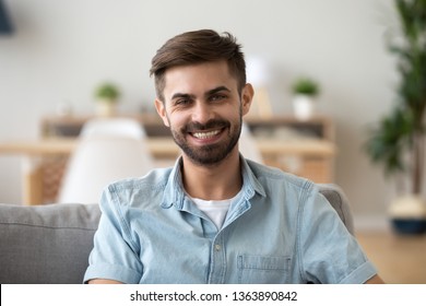 Head shot portrait of happy smiling millennial man sitting on comfortable sofa in living room, looking at camera, successful confident freelancer, excited guy posing for photo at home - Shutterstock ID 1363890842