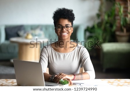 Head shot portrait of happy smiling African American woman sitting at table in cafe, looking at camera, excited female posing, working at computer, doing homework, preparing report in coffee house