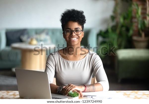 カフェのテーブルに座り カメラを見 興奮した女性のポーズを取り コンピューターで働き 宿題をし コーヒーハウスで報告書を作成するアフリカ系アメリカ人女性の幸せな笑顔のポートレート の写真素材 今すぐ編集
