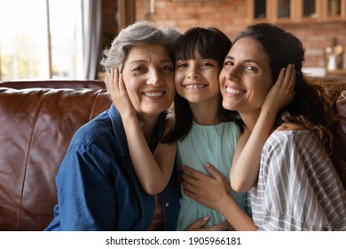 Head shot portrait happy three generations of women hugging, touching cheeks, smiling little girl sitting on couch between young mother and mature grandmother, posing for family photo at home - Powered by Shutterstock