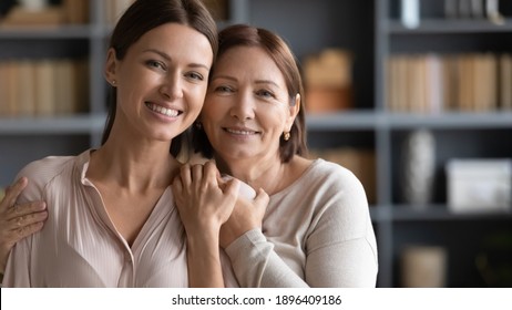 Head shot portrait happy two generations of women hugging, standing at home, smiling mature elderly mother embracing beautiful grownup daughter from back, family posing for photo together