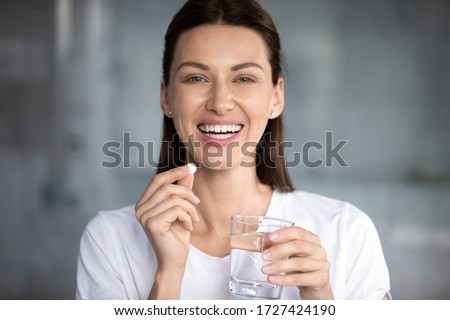 Head shot portrait happy woman holds pill glass of water looks at camera, takes daily medicine vitamin D, omega 3 supplements, skin hair nail strengthen and beauty, medication for health care concept