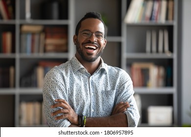 Head shot portrait laughing excited African American man wearing glasses standing with arms crossed in modern cabinet, positive young male confident businessman in eyewear looking at camera