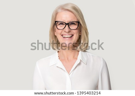 Head shot portrait laughing old businesswoman in glasses white blouse looks at camera feels happy pose isolated on grey studio background, experienced professional business coach teacher concept image