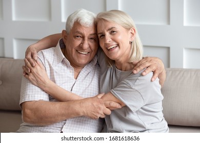 Head Shot Portrait Older Wife And Husband With Healthy Toothy Smiles Hugging, Looking At Camera, Sitting On Cozy Sofa At Home, Happy Adult Middle-aged Daughter Embracing Mature Father, Family Photo