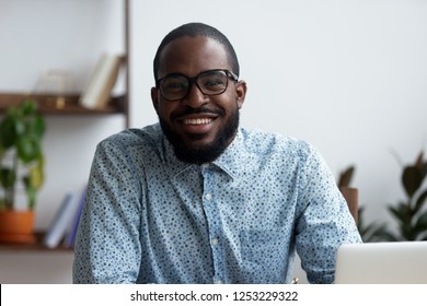 Head shot portrait of positive african american businessman in eyeglasses sitting at office desk using laptop. Confident happy with guileless smile entrepreneur looking at camera posing in workplace
