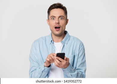 Head shot portrait shocked man looks at camera open mouth make big eyes holds phone astonished by received sms feels stunned surprised amazed pose studio grey wall, unbelievable horrified news concept