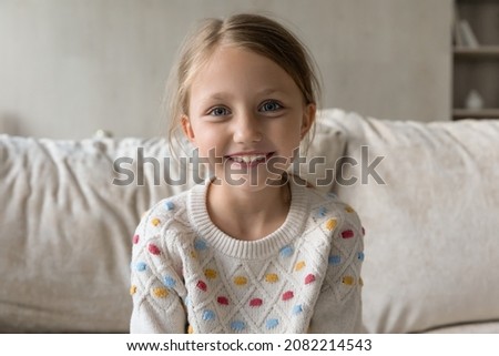 Head shot portrait of smiling adorable little kid girl sitting on comfortable couch, holding distance video call conversation, web camera view. Joyful cute small child posing alone in living room.