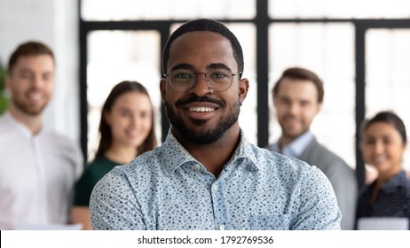 Head Shot Portrait Smiling African American Executive Wearing Glasses Standing In Modern Office Room With Diverse Employees Team, Corporate Staff Members, Successful Startup Founder Looking At Camera