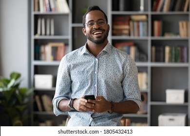 Head shot portrait smiling African American man holding phone, looking at camera, positive young businessman wearing glasses preparing to make call, using cellphone, standing in modern office room