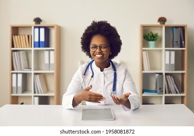 Head Shot Portrait Of Smiling African American Female Doctor Wearing Eyeglasses, Uniform With Stethoscope Speaking, Consulting Patient Online, Looking At Camera, Making Video Call Sitting At Table