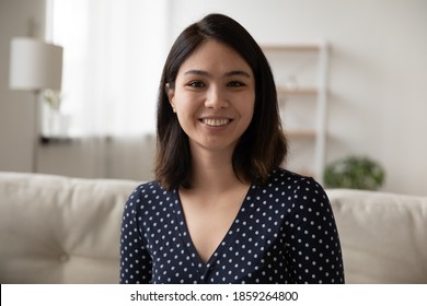Head shot portrait smiling Asian young woman looking at camera, sitting on couch, attractive female blogger shooting vlog for social network, teacher coach recording webinar, working at home