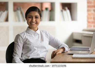Head shot portrait smiling attractive Indian businesswoman sitting at work desk in office, confident happy young woman looking at camera, posing for corporate photo, motivated student or employee