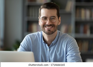 Head shot portrait smiling businessman student worker wearing glasses looking at camera, happy satisfied young man sitting at work desk with laptop, posing for photo in modern cabinet - Powered by Shutterstock