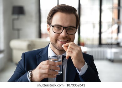 Head shot portrait smiling businessman wearing glasses taking meds, medication pills in office, holding glass of water, looking at camera, happy man employee drinking daily vitamins, supplements - Powered by Shutterstock