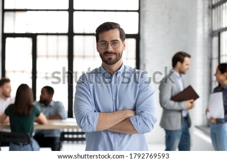 Head shot portrait smiling confident businessman wearing glasses standing in modern office room with arms crossed, diverse colleagues on background, executive boss startup founder looking at camera