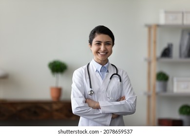 Head Shot Portrait Smiling Indian Woman Doctor With Folded Hands Standing, Confident Young Female Physician General Practitioner Therapist Wearing White Uniform With Stethoscope Looking At Camera