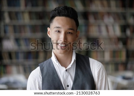 Head shot portrait of smiling Korean handsome male college student standing in modern library. Happy millennial confident 20s Asian guy with trendy hairstyle and toothy smile looking at camera.