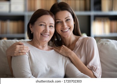 Retrato de cabeza sonriendo madre madura abrazando a hija, sentada en el sillón en casa, mirando a la cámara, joven mujer abrazando hombros de mamá más viejos, foto familiar, dos generaciones en unión Foto de stock